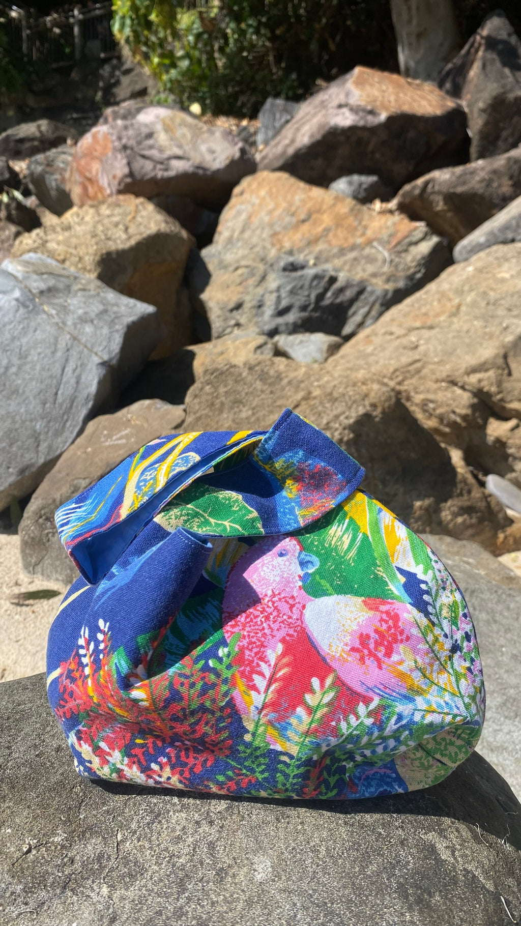 Back view of a colorful, tropical patterned fabric knot style bag with a tropical bird and foliage design, resting on a large rock outdoors with a rocky shoreline background. White branded label ISLA TIDE.