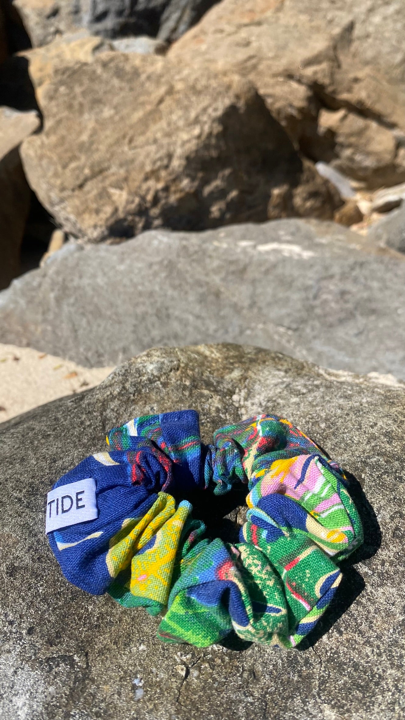 A multicolor patterned fabric scrunchie with a white label reading 'ISLA TIDE' is placed on a large gray rock outdoors, with rocks in the background.