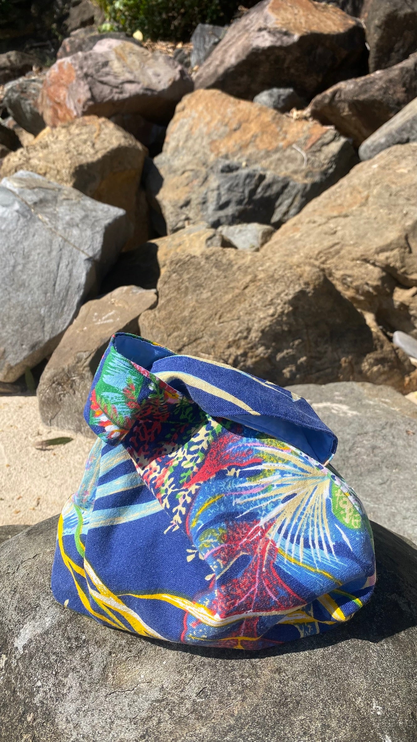 A cotton fabric Japanese knot style bag with a colorful tropical print sits on a large rock outdoors, with sunlit stones in the background. ISLA TIDE white label branding.