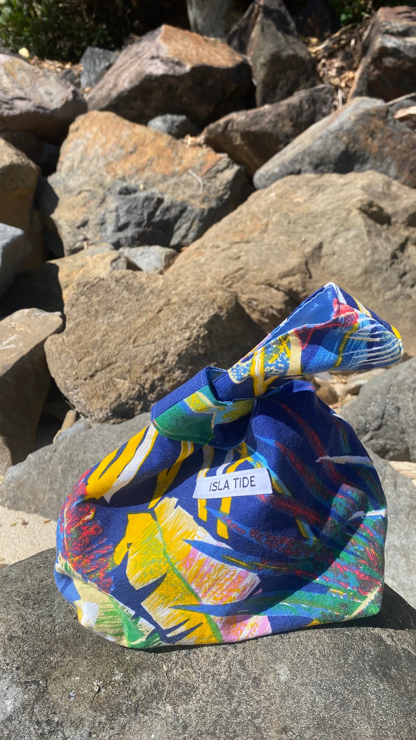 A colorful Japanese knot style fabric bag with a white branded label sits on a large rock outdoors, surrounded by stacked natural stones. The bag has a vibrant foliage tropical print in blue, yellow, green, pink, and white, with a white label reading 'ISLA TIDE'.