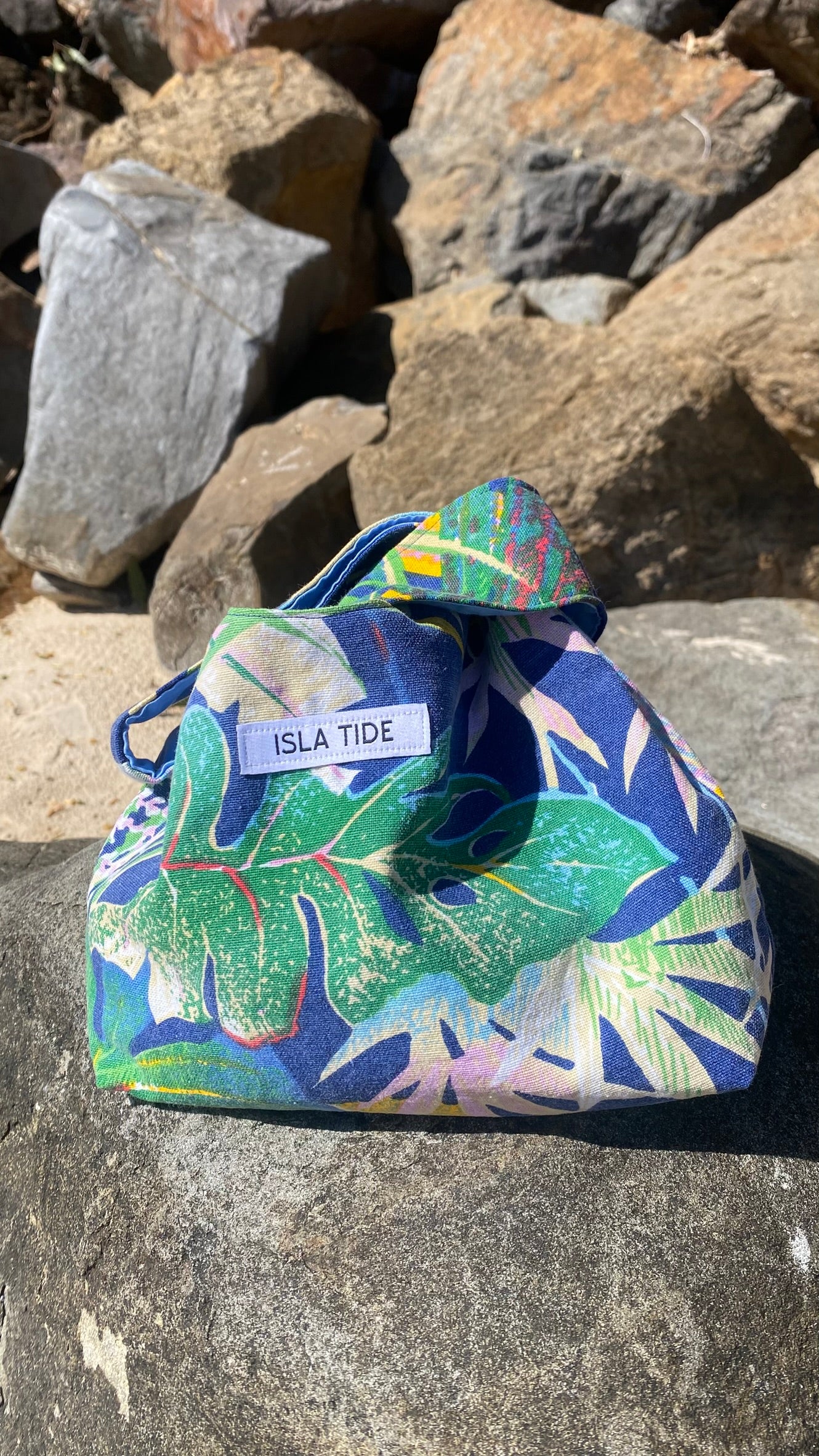 A cotton Japanese style knot bag with a tropical leaf print in blue, green, and pink, resting on a gray rock with natural rocks in the background. The pouch has a white label that reads 'ISLA TIDE'.