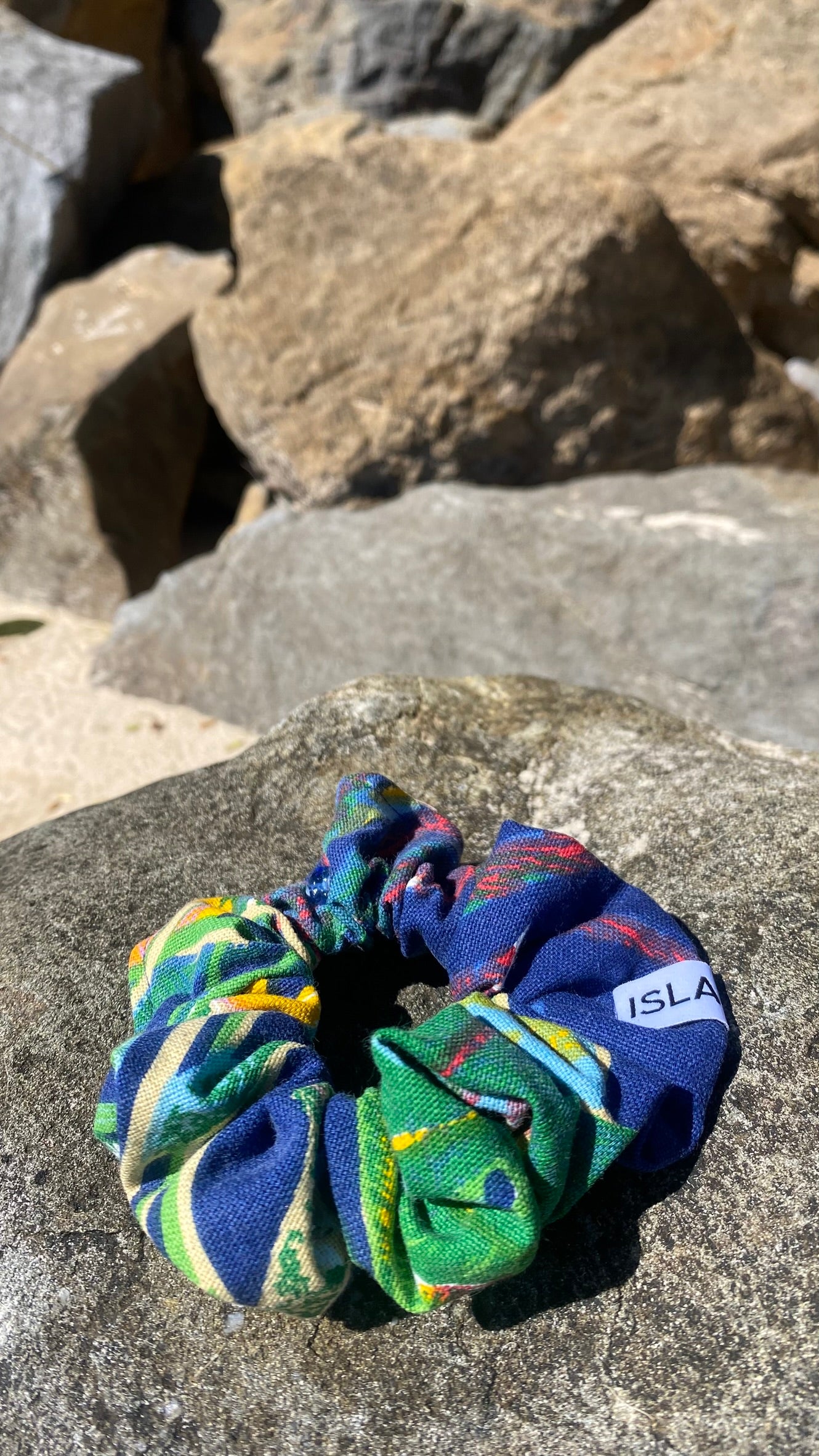 A multicolor patterned fabric scrunchie with blue, green, yellow, and red tones, resting on a gray rock outdoors with rocks in the background. A small white tag on the scrunchie reads 'ISLA TIDE'